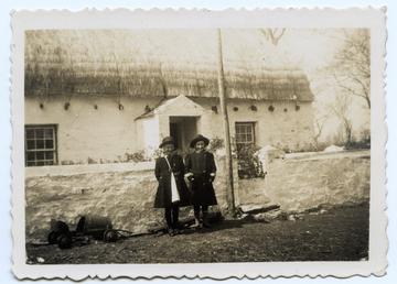 Two girls standing outside a part mud-walled cottage…